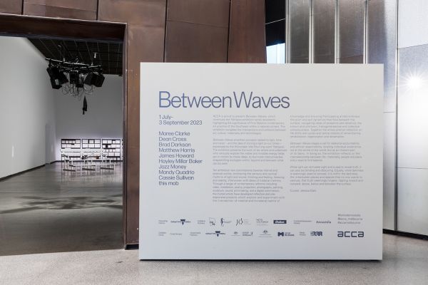 A large white wall at the entrance to a large room in an art gallery. The title wall contains the exhibition's title, Between Waves, in large text in has smaller text with information about the exhibition.