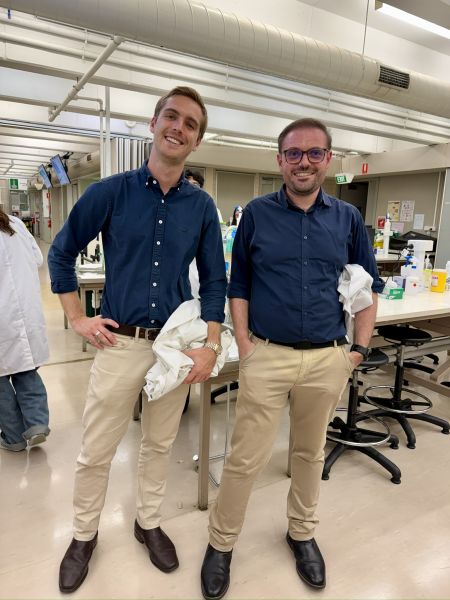Two caucasian males, Connor Daymond (fair hair and clean shaven) and Dr Chris Freelance (dark brown hair, bearded, and wearing glasses), standing in a laboratory with a white lab coat under their left arms and both wearing dark boots, tan pants, and a navy blue shirt.