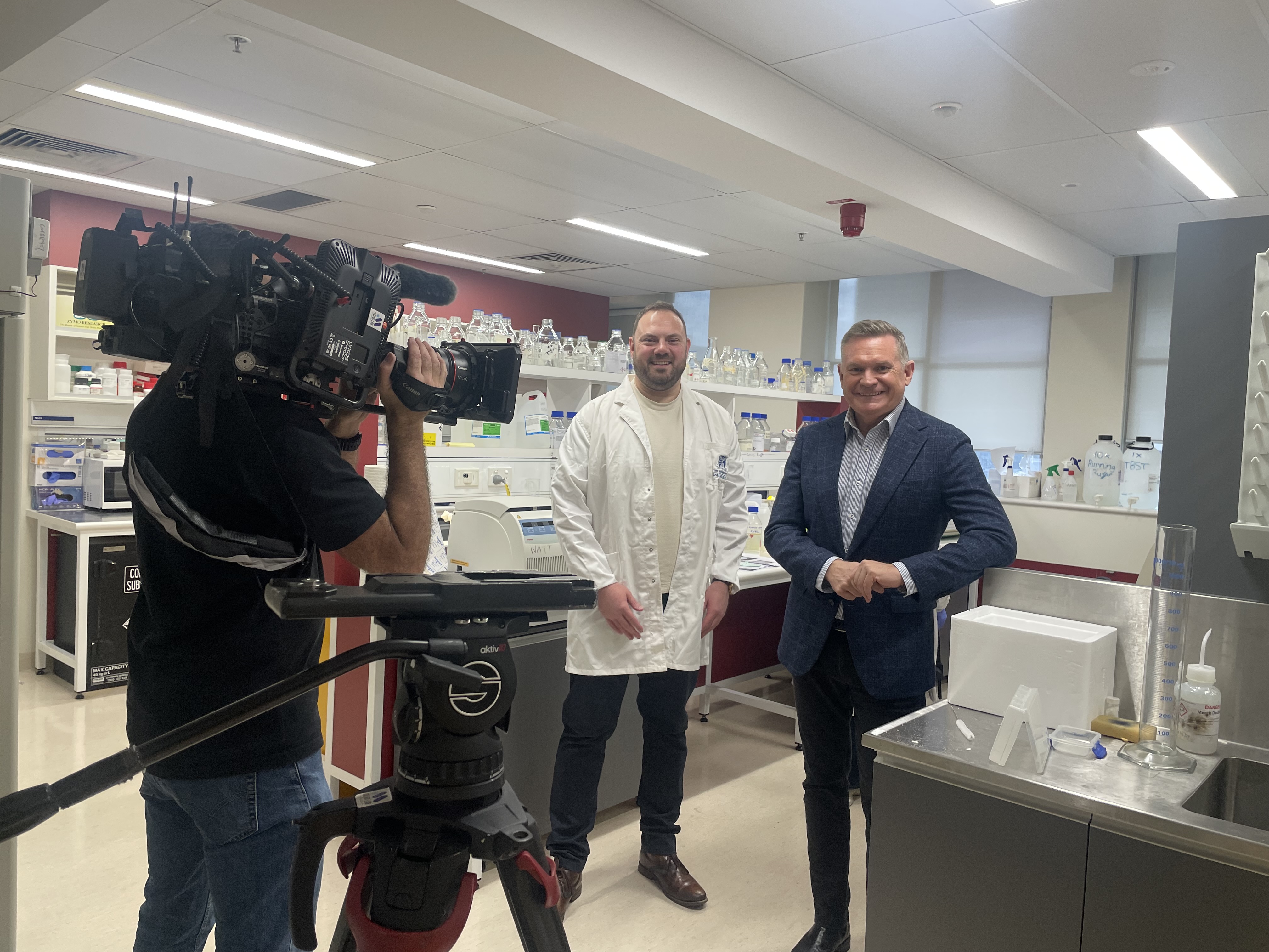 Two men stand in lab while another man holds TV camera