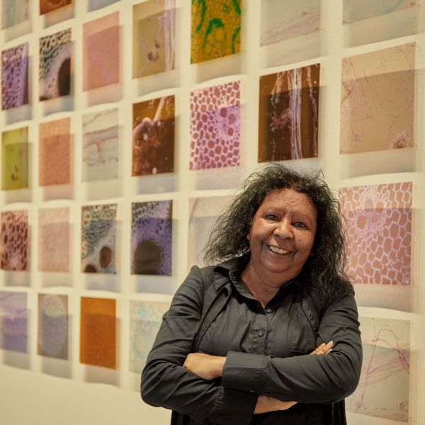 Artist Maree Clarke stands in front of a cream wall on which coloured squares are mounted. Each square is a piece of acetate on which is printed an image of a river reed stem viewed under the microscope.